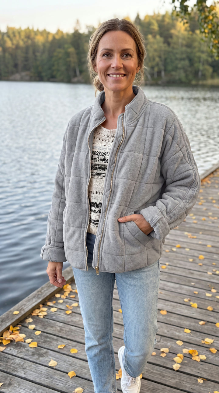 Kvinna i grå quiltad jacka med ståkrage och dragkedja, jeans, på brygga vid sjö i höstljus